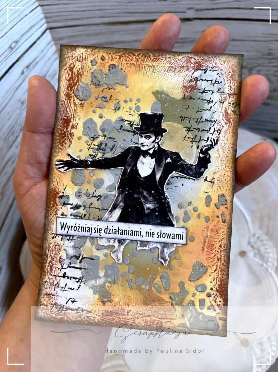 Ręcznie wykonana karta mixed‑media w stylu vintage z postacią w cylindrze i cytatem „Wyróżniaj się działaniami, nie słowami”, sfotografowana na jasnym, teksturowanym tle. Artystyczna kompozycja z warstwami farb, tekstur i detali, projekt Scrapstacja.