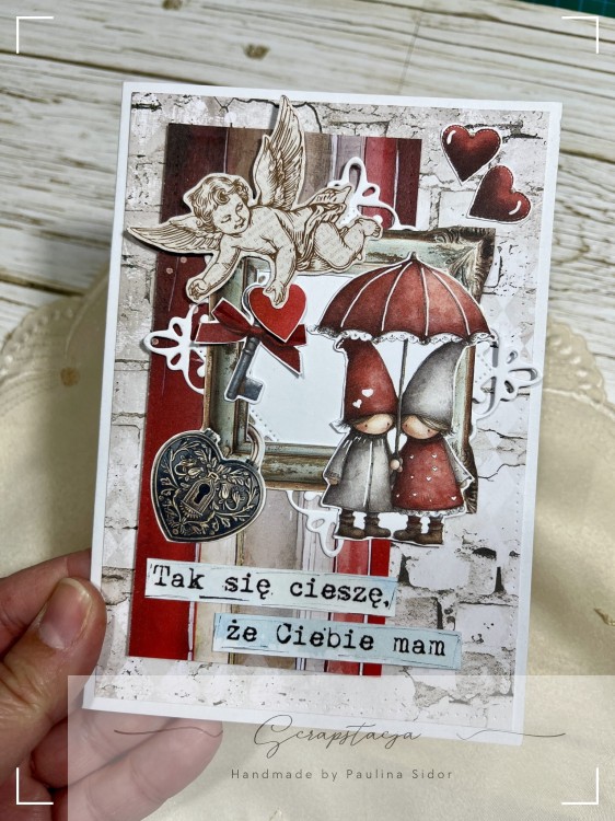 Kartka walentynkowa w stylu scrapbooking z cherubinkiem, parą pod czerwonym parasolem, sercami i dekoracjami, z napisem „Tak się cieszę, że Ciebie mam”.