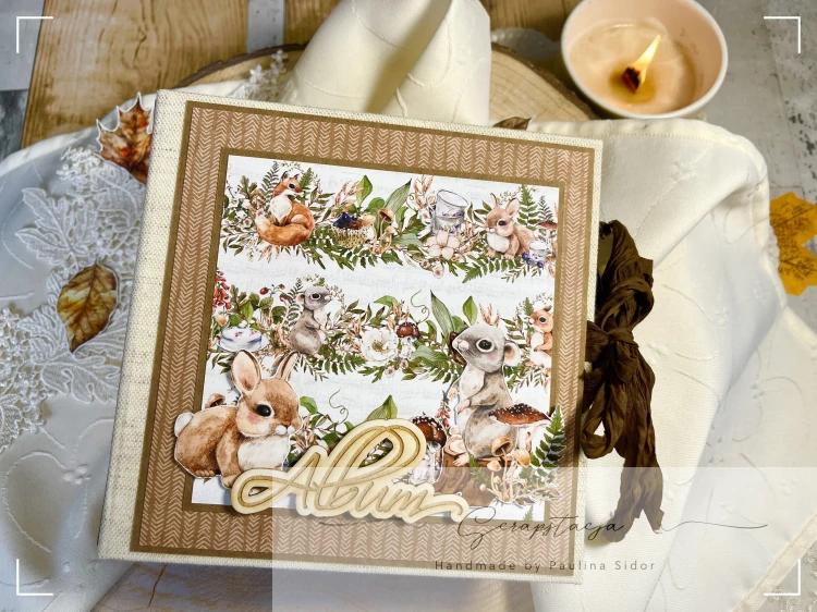 Zdjęcie przedstawia okładkę albumu wykonanego ręcznie metodą scrapbookingu w leśnym klimacie. Okładkę stanowi grafika z leśnymi zwięrzątkami i dużym napisem Album. Album zamykany na wstążkę gnieciuszkę.